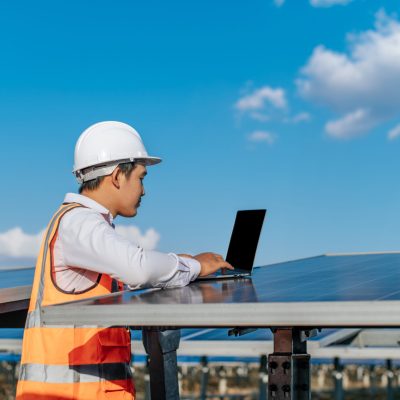 Young Asian Inspector Engineer man use laptop computer working at solar farm, Technician, supervisor male in white helmet Checking operation of sun and photovoltaic solar panel in station, copy space